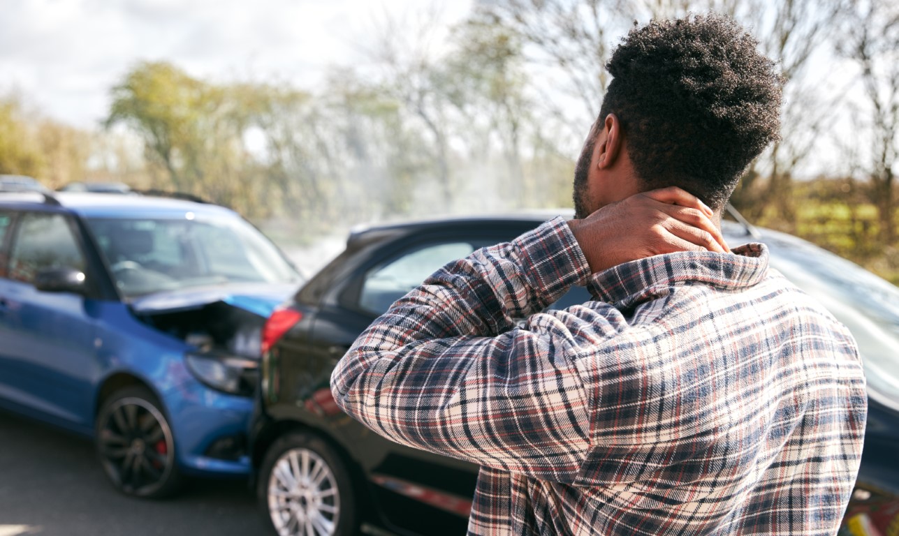 Young male driver with rear-end crash injuries standing by damaged car after road traffic accident